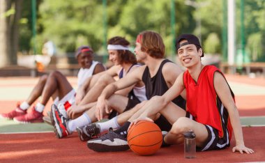 Sırıtkan Asyalı basketbolcu çeşitli takımlarıyla açık hava oyun sahasında oturuyor.