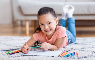 Evde yere uzanmış resim çizen sevimli küçük kız.