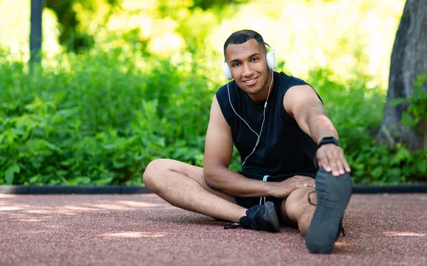 Retrato de jovem negro em fones de ouvido esticando as pernas na pista de jogging no parque, espaço cópia — Fotografia de Stock