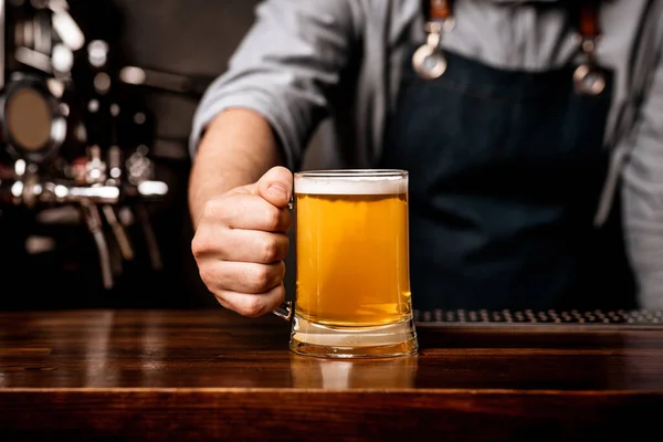 Barman en delantal sirve cerveza en taza de vidrio en barra de madera contador —  Fotos de Stock