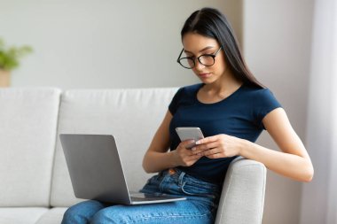 Evde koltukta oturan dizüstü bilgisayarlı kız cep telefonu kullanıyor.