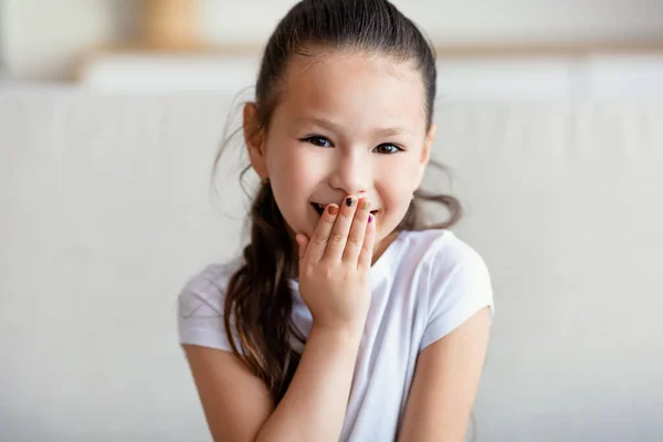 Carino ragazza ridente copertura bocca seduta sul divano a casa — Foto Stock