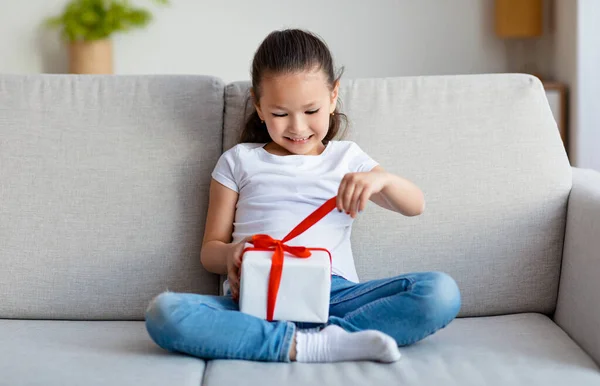 Happy Asian Girl Opening Gift Box Sitting On Couch Indoors — Stok Foto
