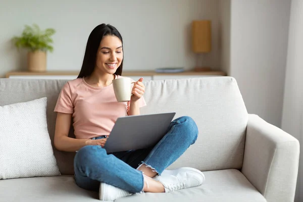 Mujer feliz usando el ordenador portátil disfrutando de café sentado en el sofá interior —  Fotos de Stock