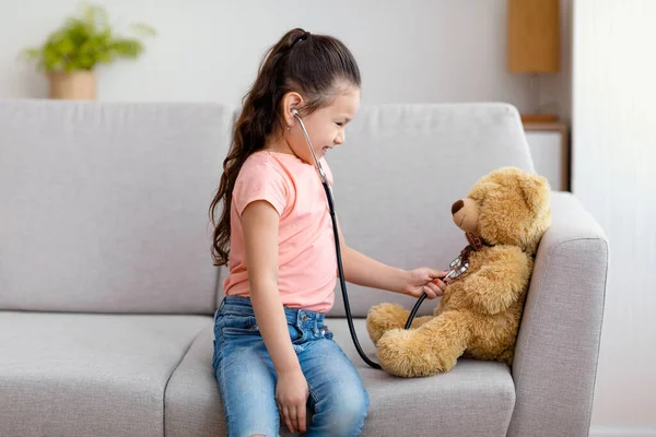 Menina jogando médico com ursinho de pelúcia sentado no sofá dentro de casa — Fotografia de Stock