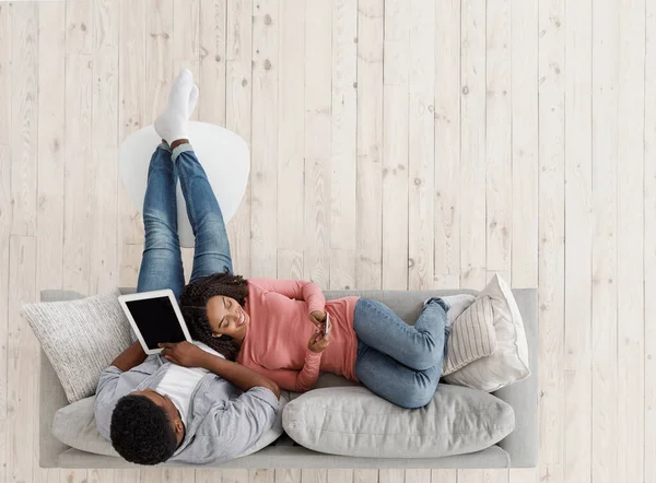 Lazer de fim de semana. Casal preto relaxante no sofá com tablet digital e smartphone — Fotografia de Stock