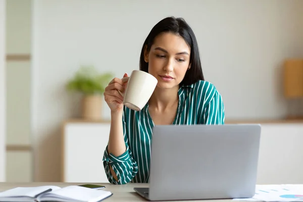 Escritório menina no laptop tomando café sentado no local de trabalho dentro de casa — Fotografia de Stock