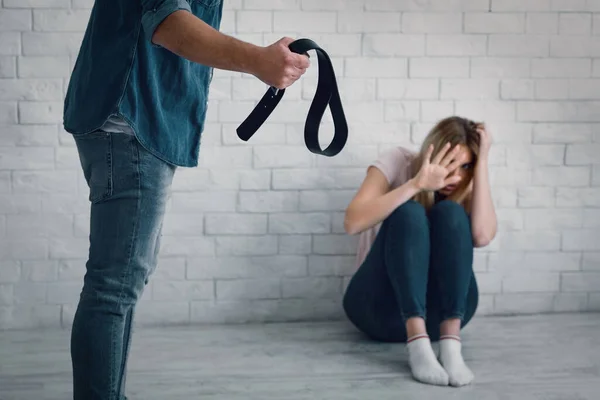 Victim and domestic violence. Aggressive man waving belt at wife sitting on floor — Stock Photo, Image