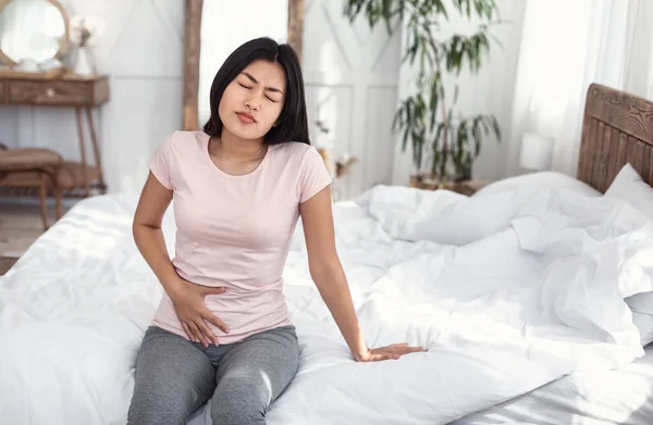 Japanese Girl Touching Aching Lower Abdomen Sitting On Bed Indoors — Stock Image Japanese Girl Touching Aching Lower Abdomen Sitting On Bed Indoors Stock Image