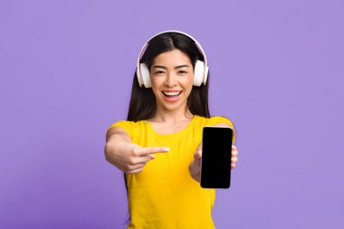 Müzik uygulaması. Siyah akıllı telefon ekranını işaret eden Haadphone 'lu neşeli Asyalı kız