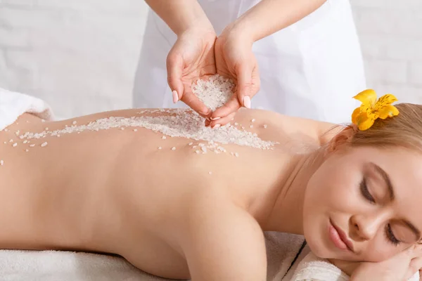 Manos de profesional haciendo masaje con sal para mujer con los ojos cerrados en la mesa —  Fotos de Stock
