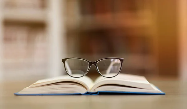 Geopend boek met bril, boekenplanken op de achtergrond — Stockfoto