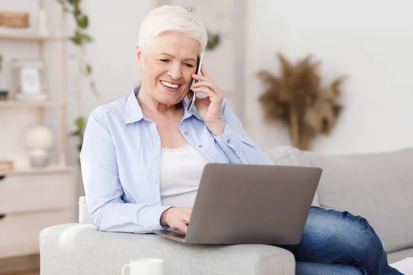 Nowoczesna starsza pani pracująca na laptopie i rozmawiająca przez komórkę w domu — Zdjęcie stockowe