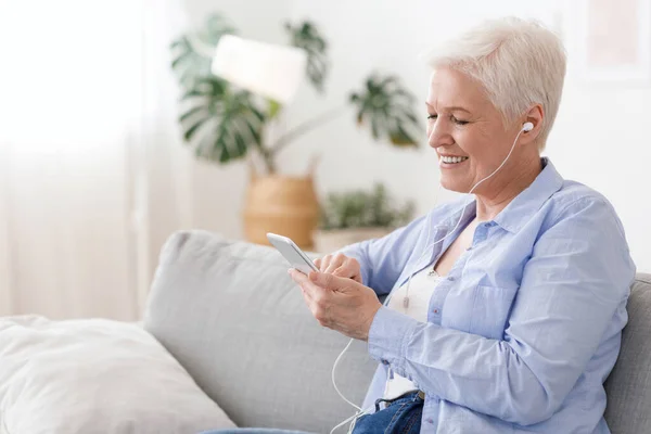 Dama de edad avanzada moderna disfrutando escuchando música en el teléfono inteligente en casa, usando auriculares — Foto de Stock