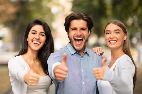 Tres alegres amigos sugiriendo pulgares hacia arriba aprobando algo parado afuera — Foto de Stock