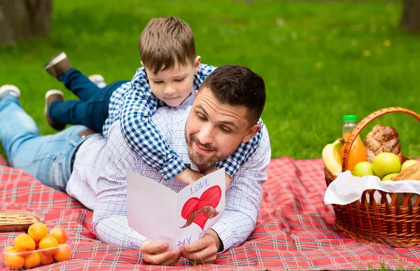 Celebración del Día del Padre. Alegre papá leyendo sus hijos tarjeta de felicitación en el picnic en el parque público — Foto de Stock