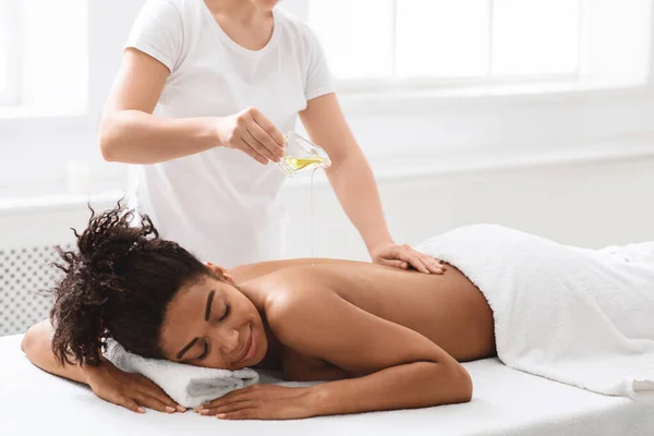 Terapeuta vertiendo aceite de masaje en la espalda dama africana —  Fotos de Stock