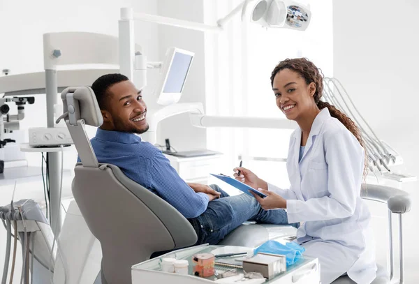 Dentista sorrindo feminino conversando com paciente, preenchendo formulário médico — Fotografia de Stock