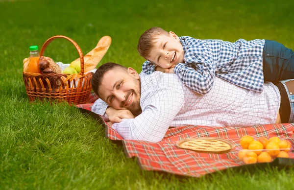 Sonriente papá con su pequeño hijo acostado en manta a cuadros juntos durante el picnic — Foto de Stock