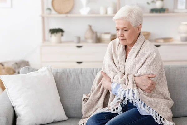 Senior senhora sentada no sofá coberto com cobertor, tremendo com alta temperatura — Fotografia de Stock
