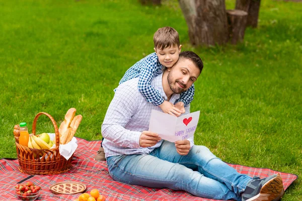 Sorpresa para papá. Sonriente papá leyendo padres día tarjeta hecha a mano de su hijo durante el picnic en el bosque — Foto de Stock