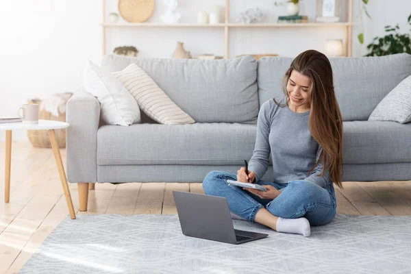 Glückliche Frau, die auf dem Boden sitzt, Notizen macht, Laptop benutzt — Stockfoto
