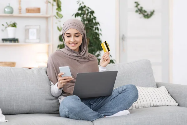 Pagos en línea. Chica árabe feliz usando teléfono inteligente, portátil y tarjeta en casa — Foto de Stock