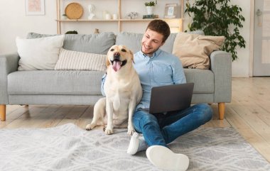 Evde dizüstü bilgisayarı ve mutlu köpeği olan genç bir adam var.