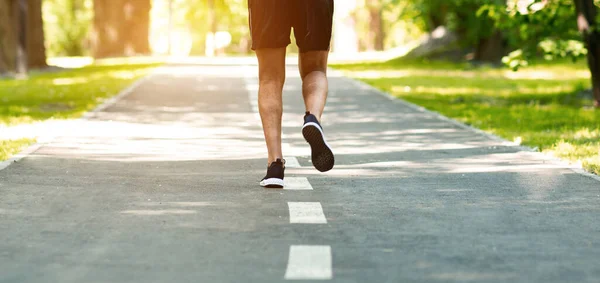 Irreconocible millennial chico corriendo a lo largo de asfalto carretera en el parque urbano, primer plano de las piernas — Foto de Stock