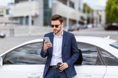 Çekici iş adamı şehir caddesinde arabasının yanındaki cep telefonundan e-postaları kontrol ediyor.