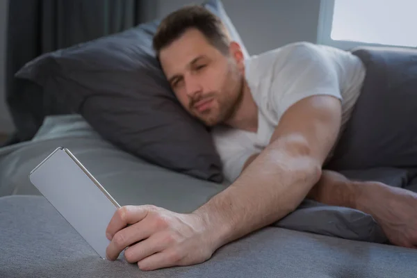 Mężczyzna za pomocą smartfona budzi się leżąc w łóżku w domu — Zdjęcie stockowe