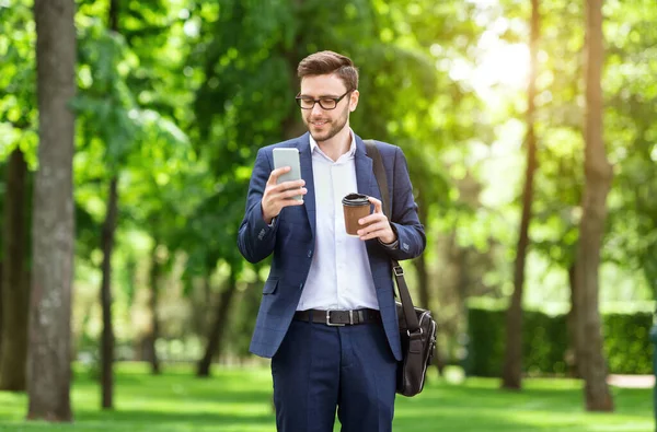 Attractive entrepreneur with coffee checking emails on smartphone at park on sunny summer day — Stock Photo, Image