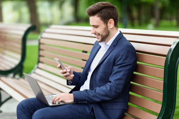 Empresário bem sucedido trabalhando on-line usando computador portátil e smartphone no banco no parque — Fotografia de Stock