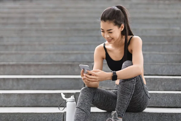 Depois do treino. Sorrindo asiático menina sentado em escadas ao ar livre e usando smartphone — Fotografia de Stock