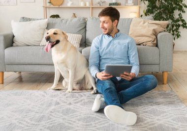 Evde elinde tablet ve labrador olan genç bir adam var.
