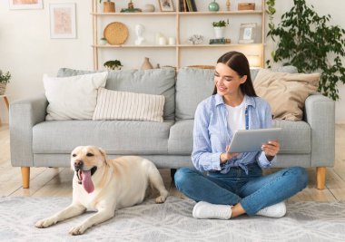 Evde elinde tablet ve labrador olan genç bir kadın var.
