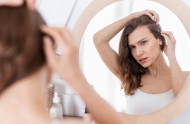 Young woman checking for thinning hair in mirror at home clipart
