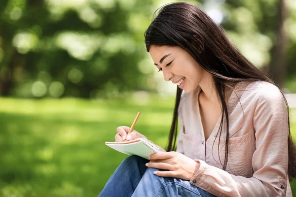 Молода азіатська студентка сидить на газоні в парку і пише нотатки — стокове фото