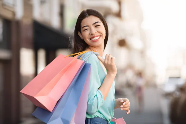 Sonriendo mujer asiática llevando bolsas de compras al aire libre y posando a la cámara — Foto de Stock
