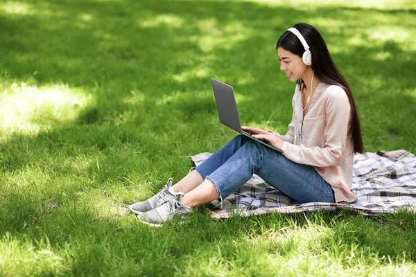 Усміхнена азіатська дівчина розслабляється з ноутбуком і навушниками на газоні в парку — стокове фото