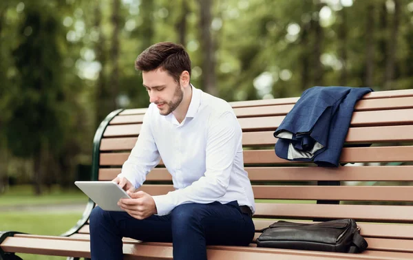 Empregado da empresa atraente relaxando no banco no parque e navegando na internet no computador tablet — Fotografia de Stock