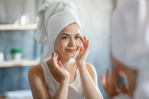 Mujer encantadora con toalla de baño en el cabello tocando su piel brillante cerca del espejo en casa — Foto de Stock