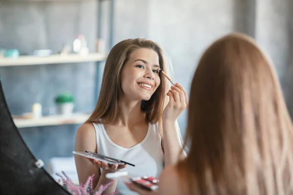 Huismake-up routine. Lieve dame aanbrengen oogschaduw op haar gezicht in de buurt van spiegel in de badkamer — Stockfoto