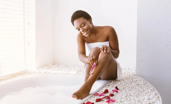 Hari kecantikan. Senyum african american girl menghilangkan rambut dengan pisau cukur, duduk di dekat mandi — Stok Foto