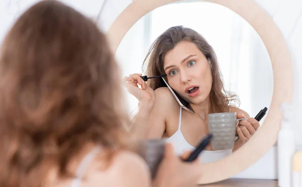 Shocked young woman talking on phone while putting mascara — Stock Photo, Image