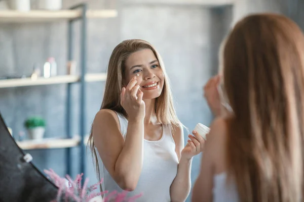 Γοητευτική κυρία εφαρμογή κάτω από την κρέμα προσώπου μάτι στο όμορφο δέρμα της κοντά στον καθρέφτη στο μπάνιο — Φωτογραφία Αρχείου