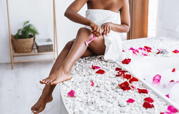 Opieka osobista w domu. Dziewczyna w białym ręczniku goli nogi, siedząc na krawędzi w łazience z pianką i płatkami róż — Zdjęcie stockowe