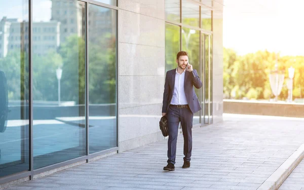 Corporate employee talking on mobile phone while walking near office building at city centre, blank space — Stock Photo, Image