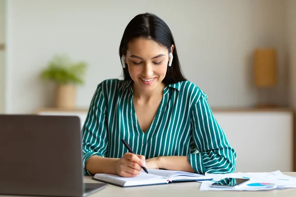 Geschäftsfrau, die einen Anruf erhält, trägt Ohrhörer und macht sich Notizen am Arbeitsplatz — Stockfoto