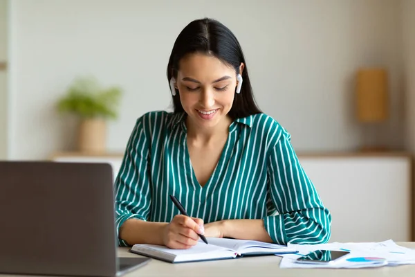 Imprenditrice che riceve chiamate indossando auricolari e prendendo appunti sul posto di lavoro — Foto Stock